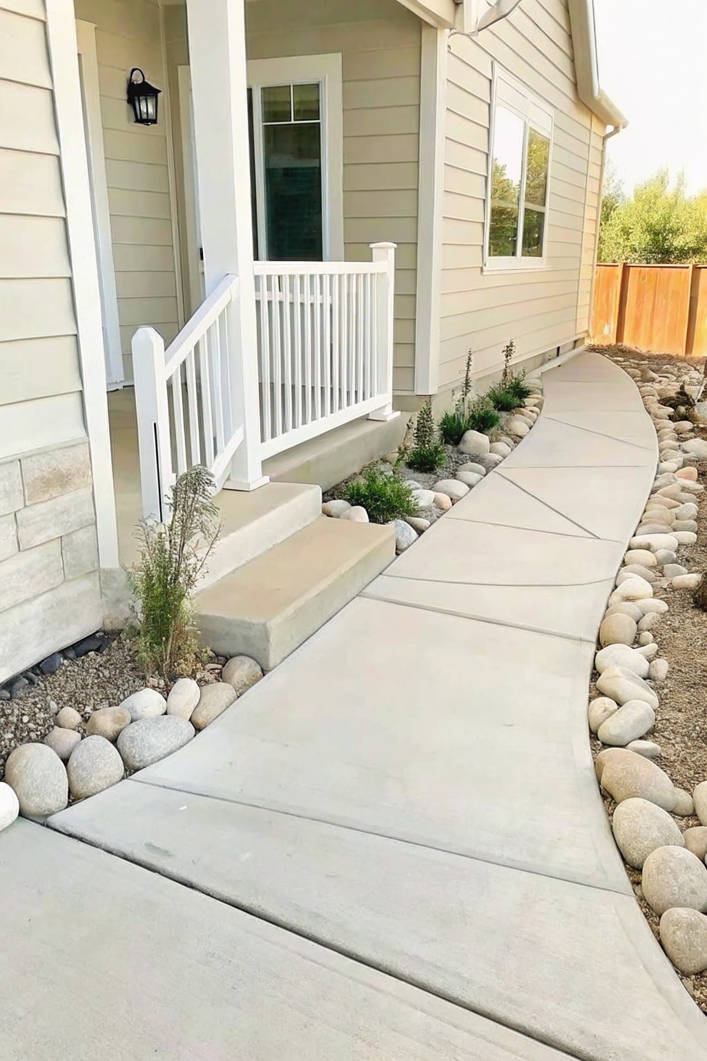 A concrete walkway curves alongside a house, bordered by smooth river rocks that edge a narrow flower bed with low green plants.