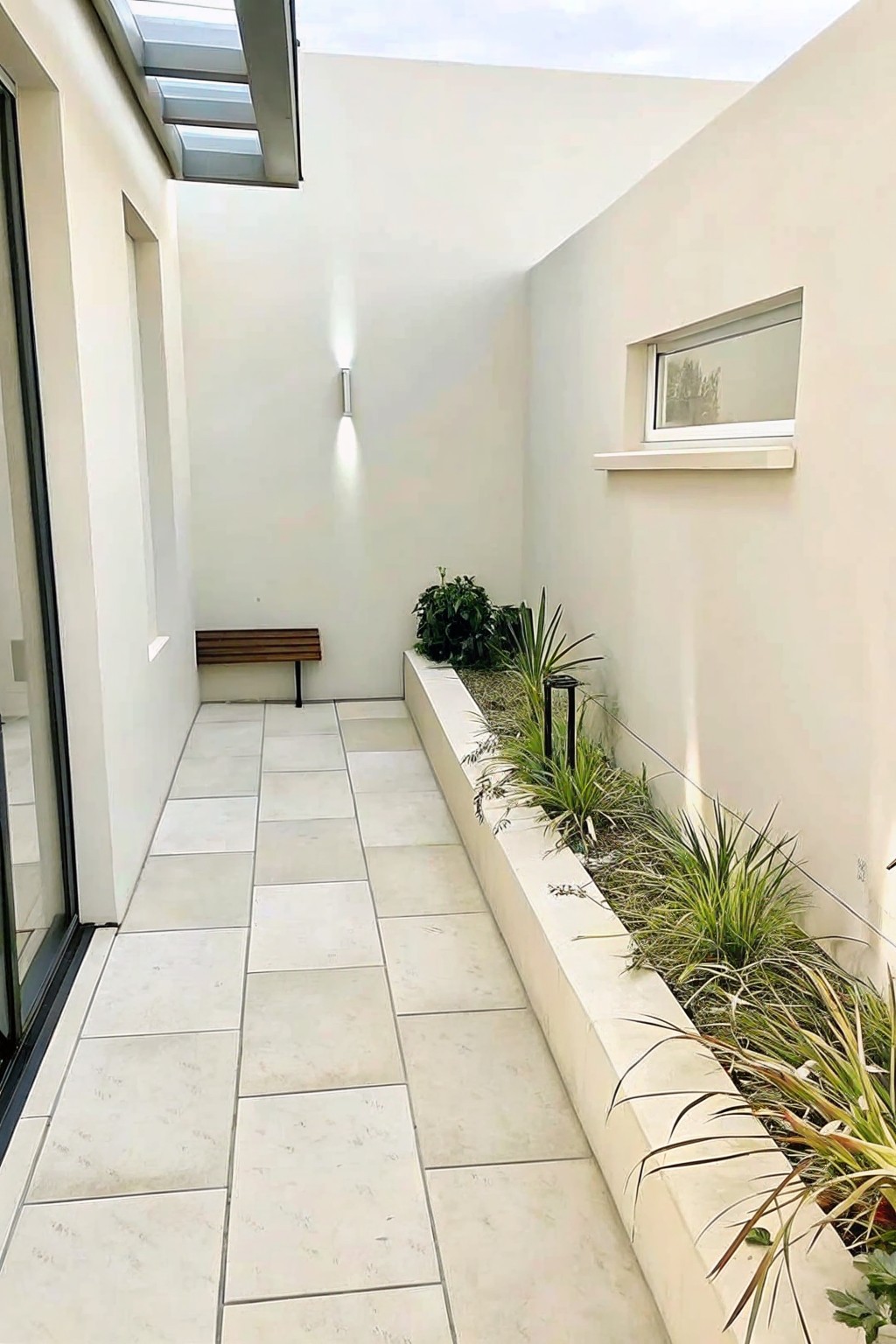 A narrow tiled walkway runs beside a long raised concrete planter filled with dense ornamental grasses and shrubs against white walls.