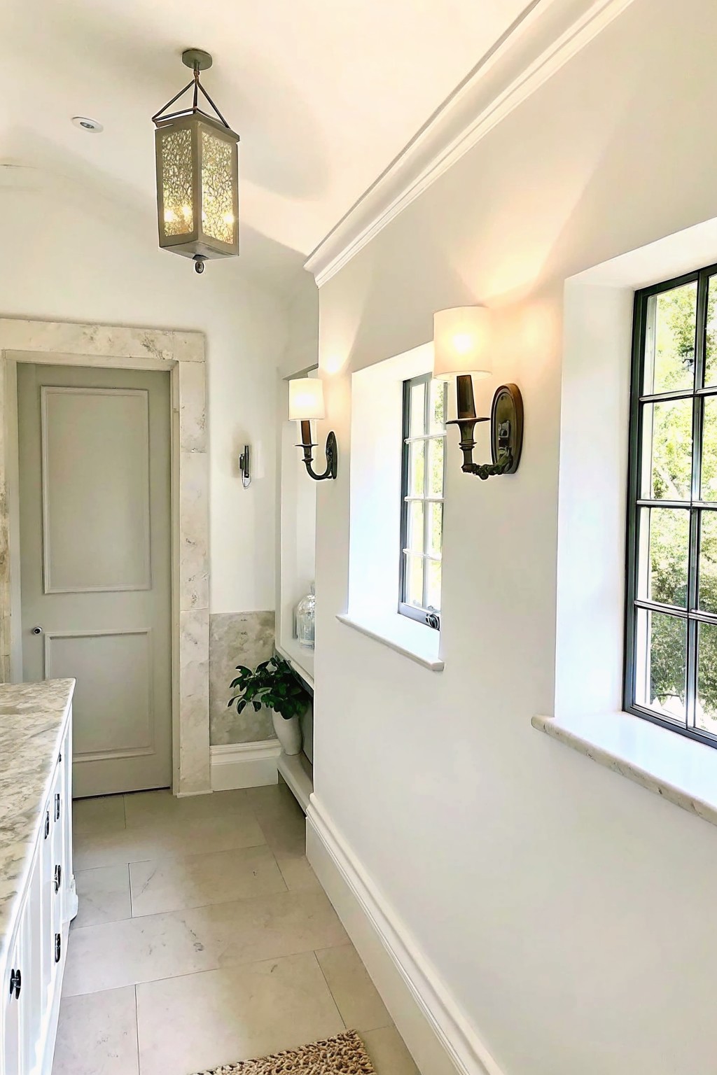 Interior hallway view showing a gray door framed by white marble trim, wall sconces, a hanging lantern, and windows with stone sills.