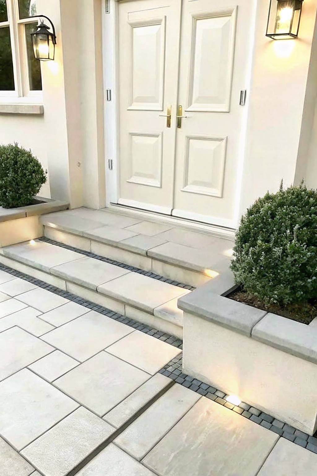 White double doors above stone steps with mosaic accents, shrubs, and glowing lanterns.