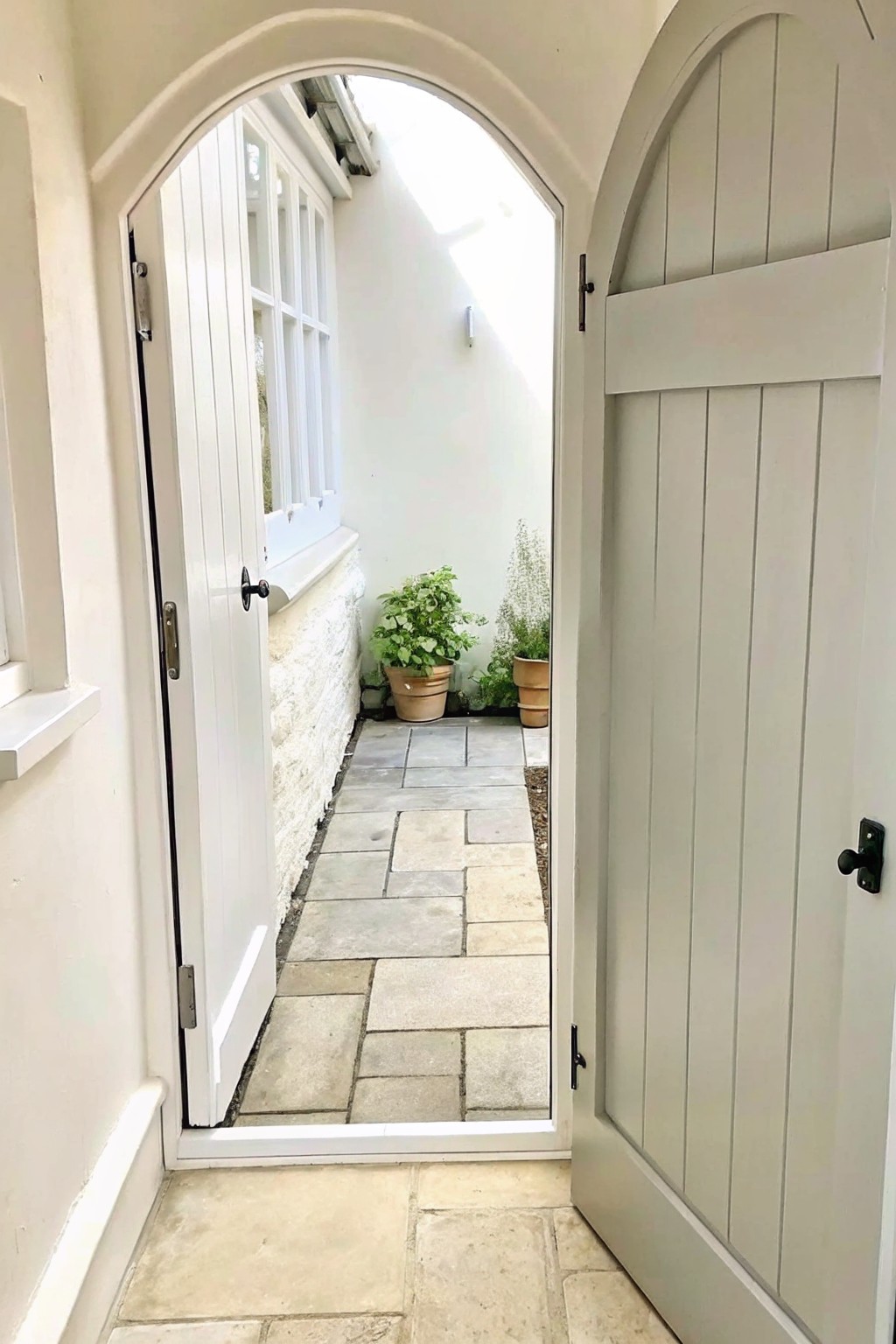 A narrow stone pathway leads through an arched opening between two white doors, with several terracotta pots holding green plants placed along the edges.