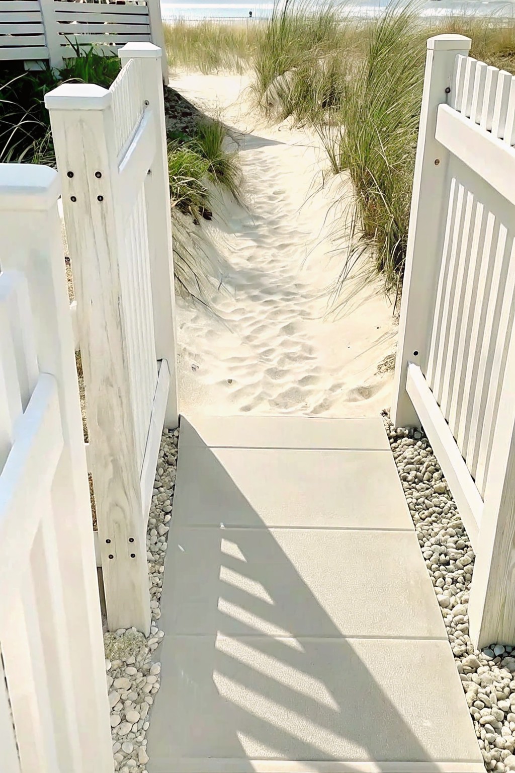 Light stone steps bordered by gravel lead to a sandy path with white fence posts on both sides and tall dune grasses.