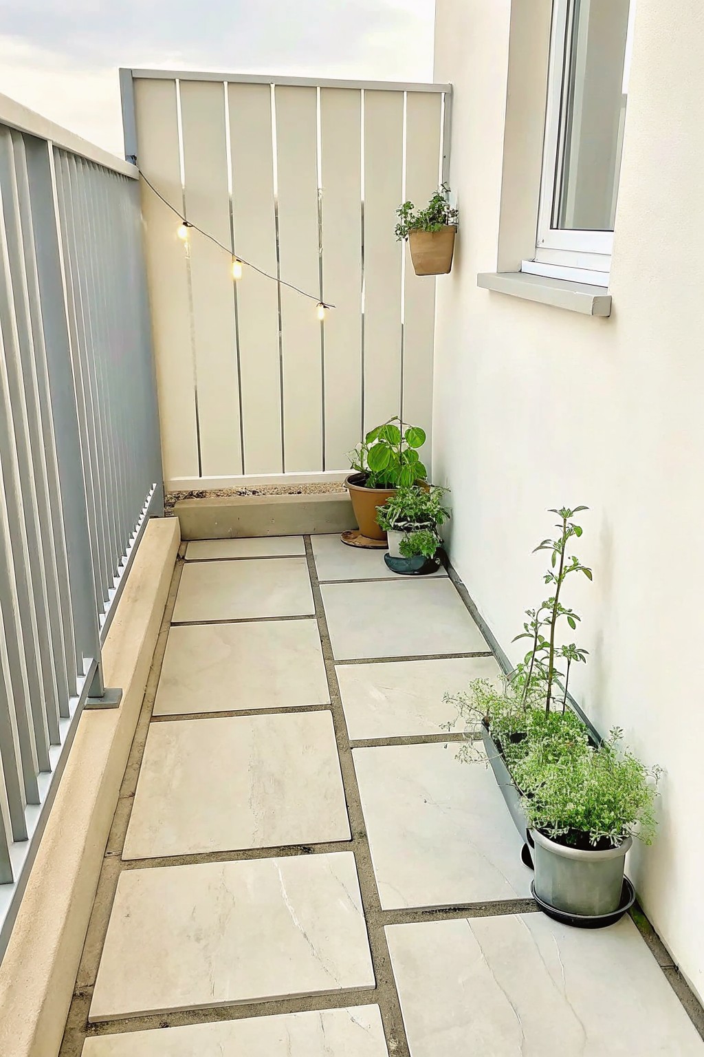 Narrow balcony with light tiles, potted plants, string lights, and white walls.