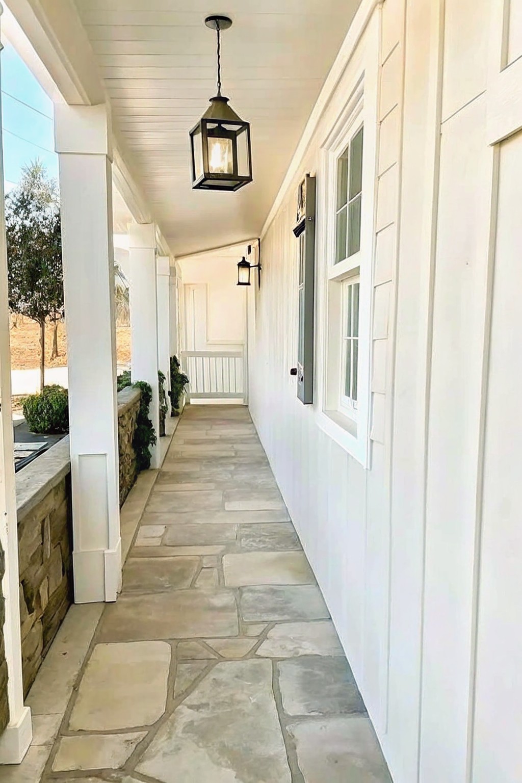 A covered walkway with irregular flagstone flooring runs alongside a white house with windows and a hanging lantern.