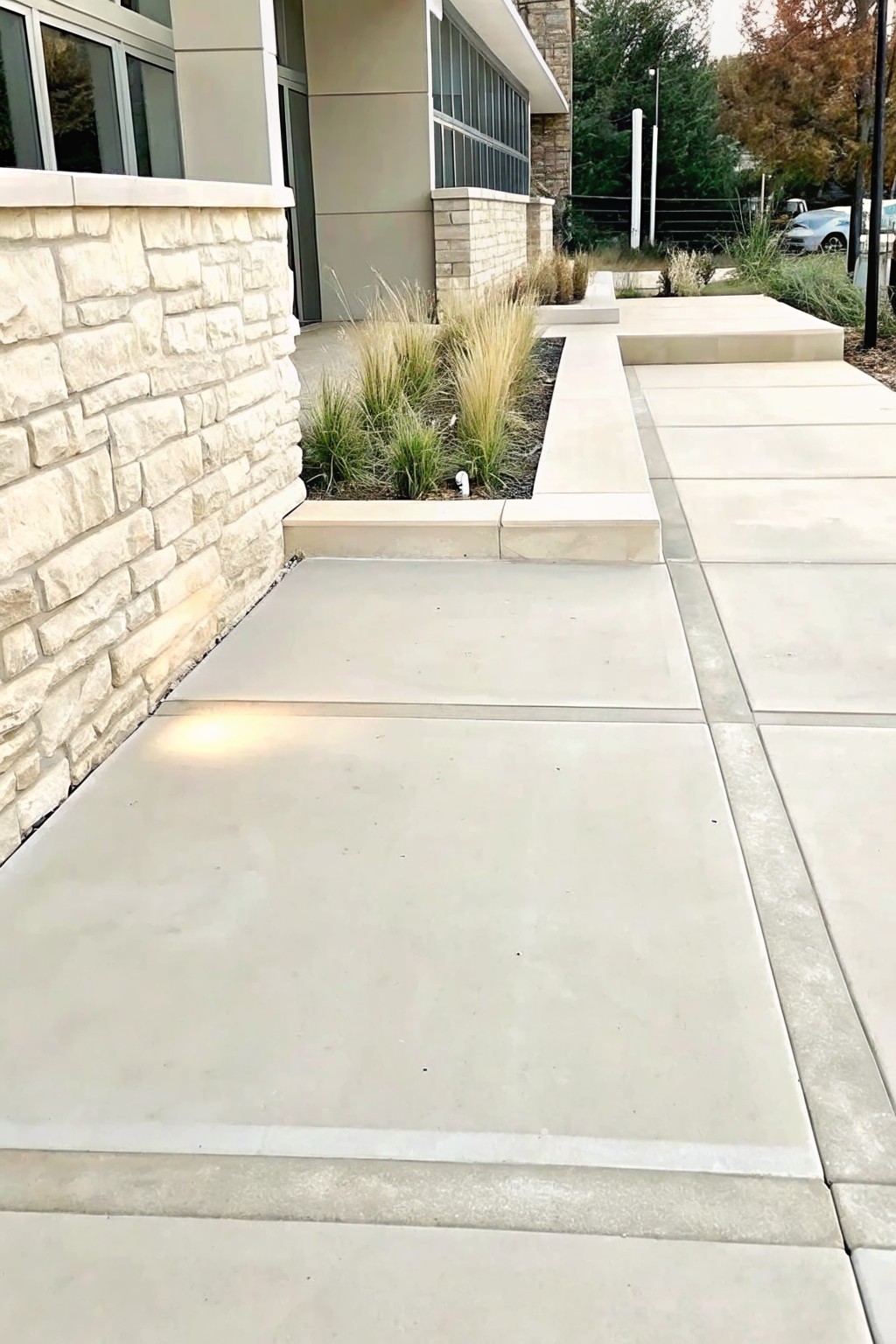 Concrete walkway beside stone wall leads to modern building entrance with grass beds.