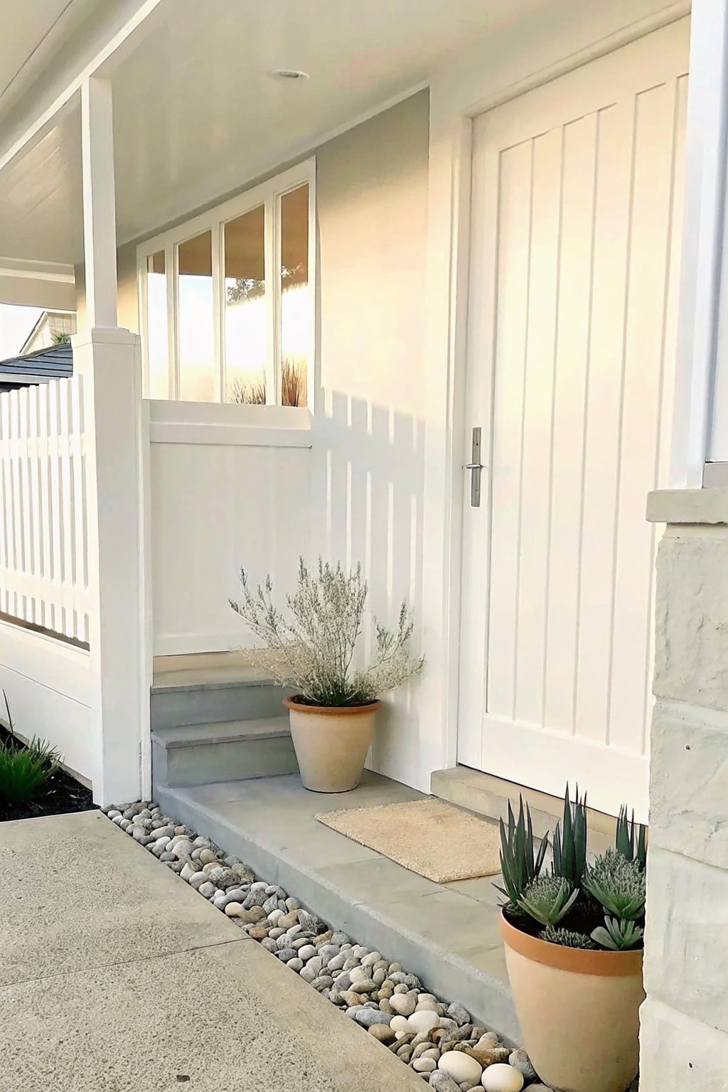 Concrete front steps with a pebble border along the edge, a white door to the right, and two potted plants on the landing.