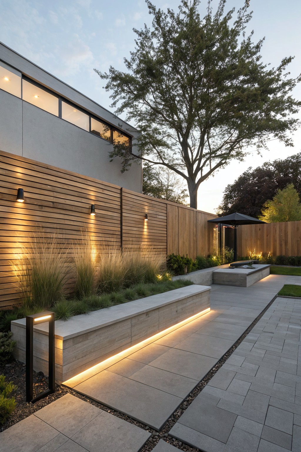 Modern backyard patio with wooden fence, illuminated concrete benches, stone tiles, and large tree at dusk.