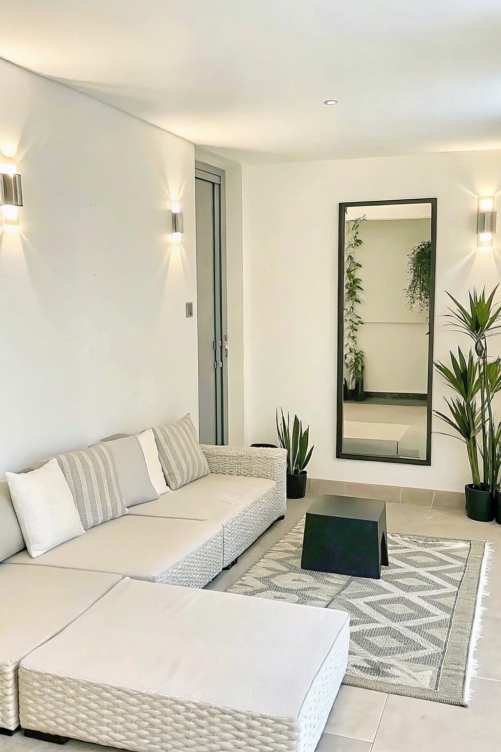 A modern outdoor patio features a white modular sectional sofa on a patterned rug, a black coffee table, several tall potted plants, and a large wall mirror.