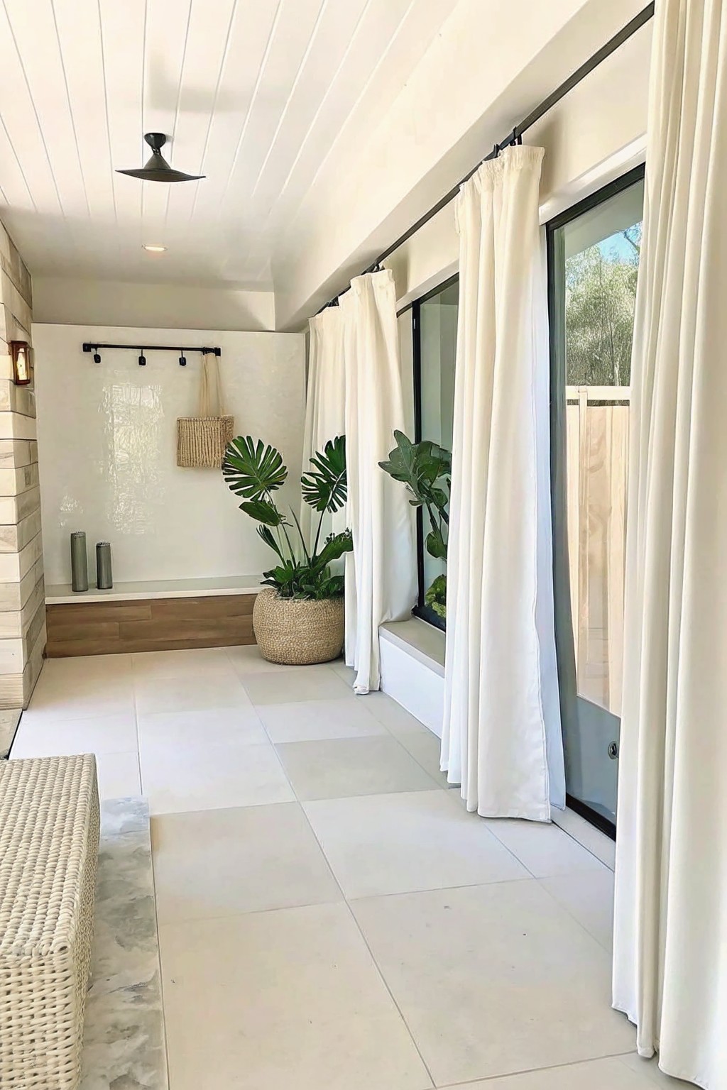 Modern indoor space with white tiled floor, large potted plants in woven containers, white curtains on a black track, and a woven bench along the left wall.