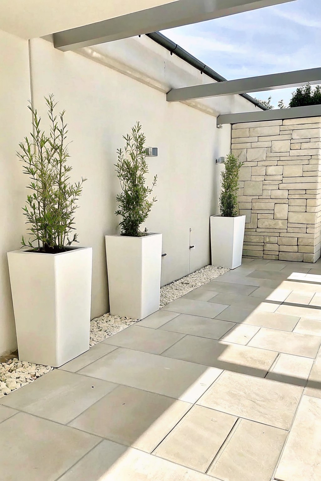 Three tall white rectangular planters with green plants stand along a white wall on a paved patio beside a stone accent wall.