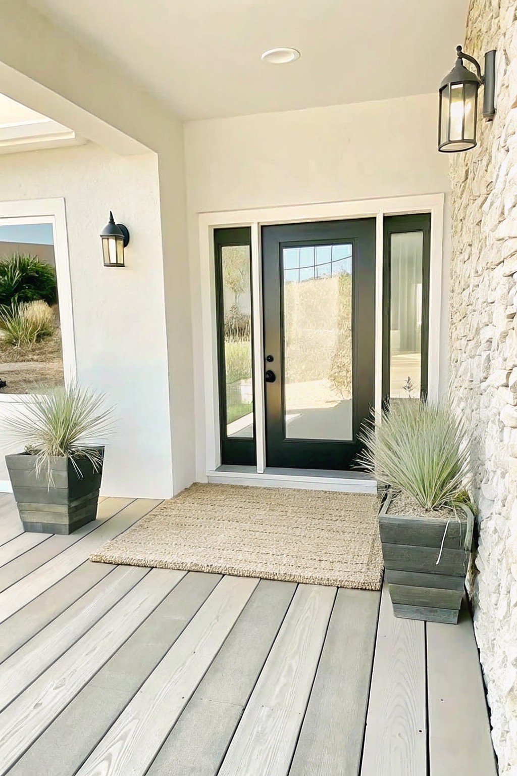 Modern home entry porch with light gray decking, a beige woven rug, dark front door with sidelights, and two large dark gray rectangular pots holding tall ornamental grasses.
