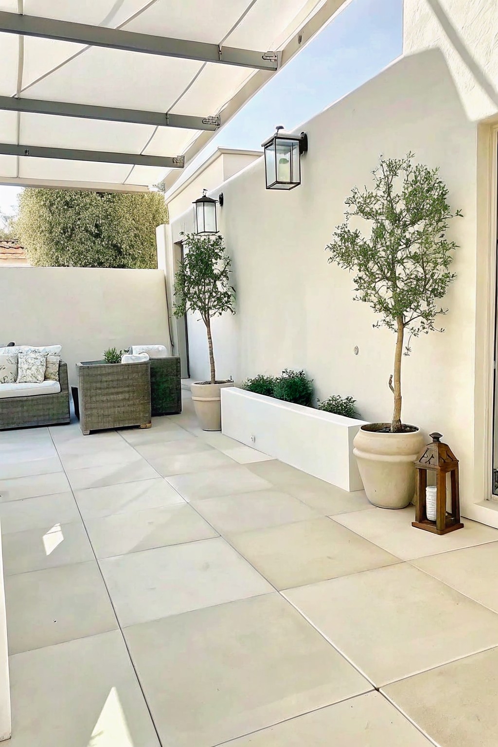 Modern patio with beige tiles, potted trees, outdoor sofa, and wall lanterns