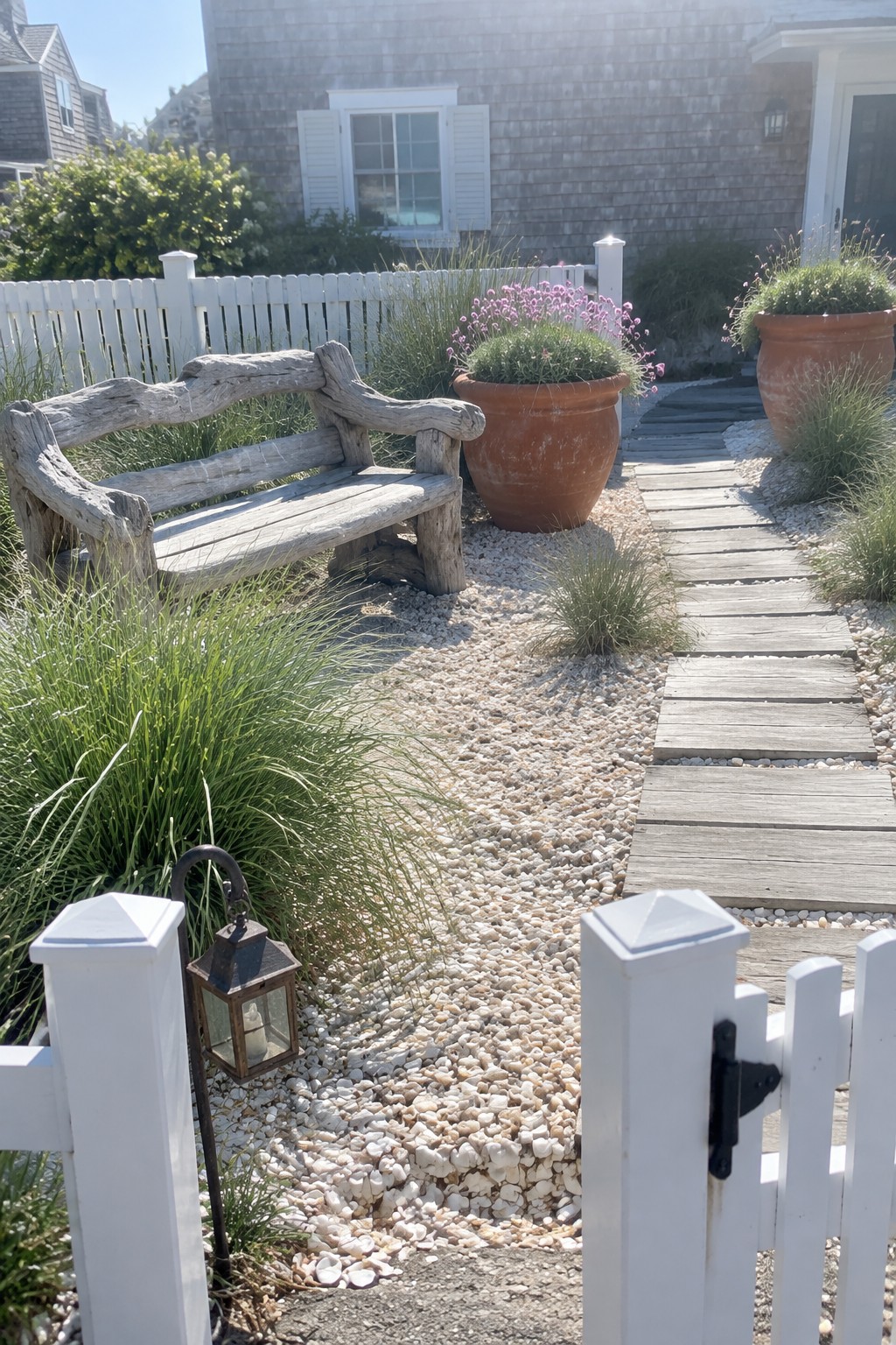 A gravel garden path with a rustic wooden bench, terracotta pots, and a wooden plank walkway leading toward a shingled house.