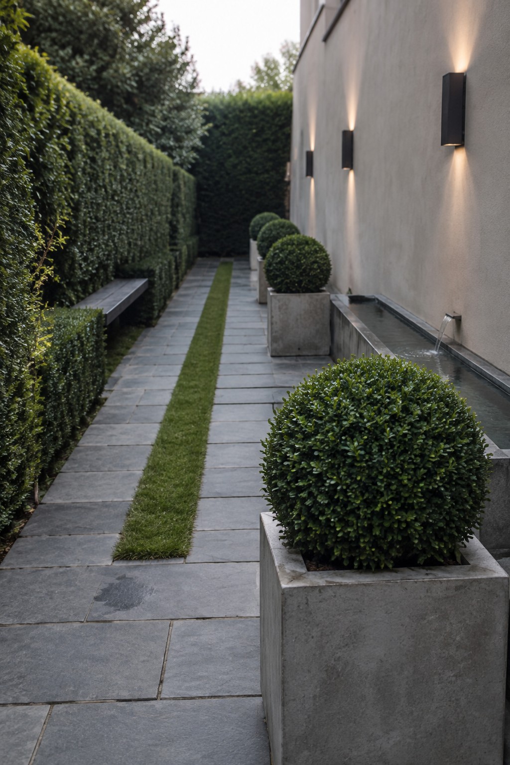Stone path with grass strip, topiary planters, wall lights and water feature.