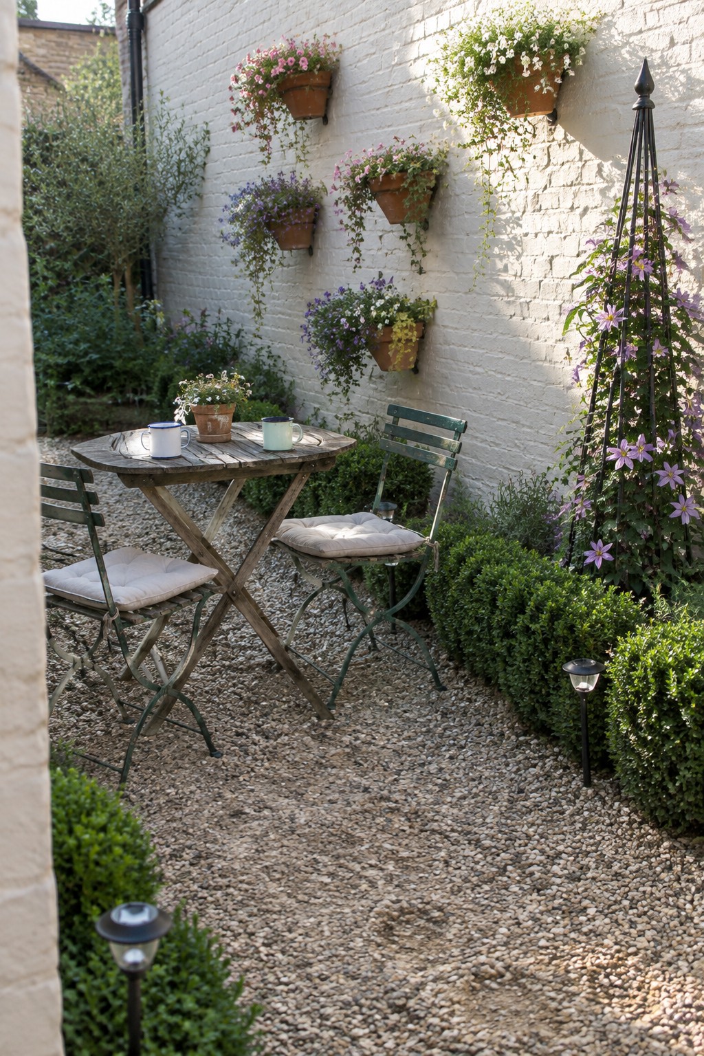 A gravel backyard patio with a wooden table and two chairs in front of a white brick wall lined with hanging terracotta pots and a tall metal trellis covered in purple flowers.