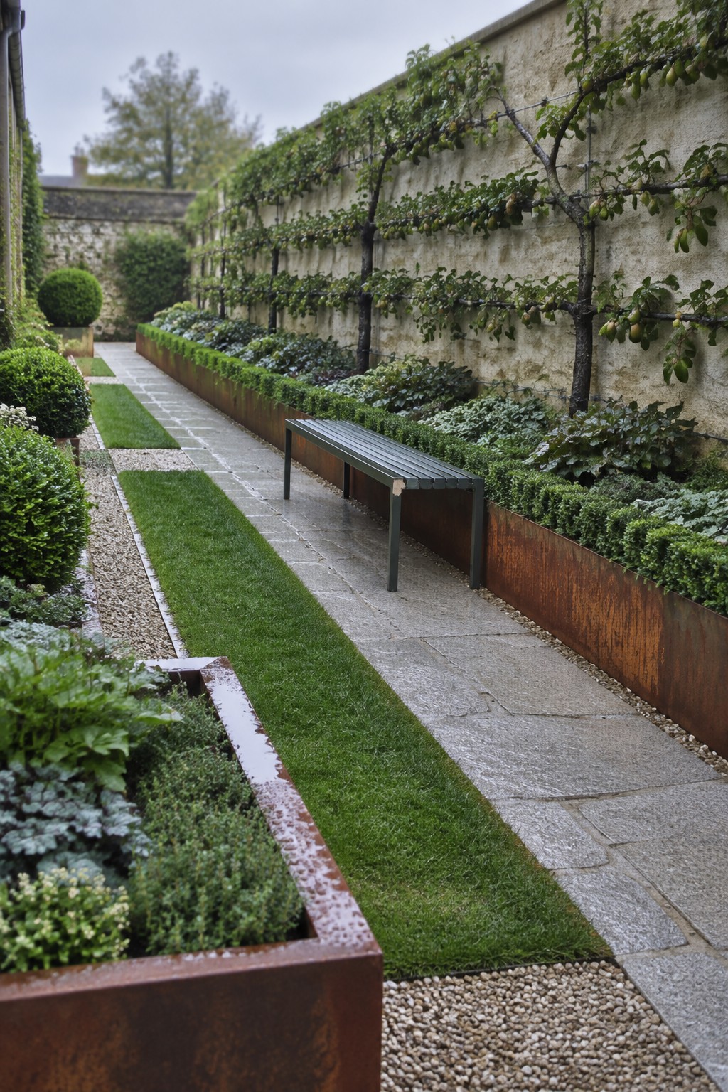 A long formal garden path lined with stone pavers, grass strips, gravel, and raised metal planting beds beside a wall with espaliered trees.