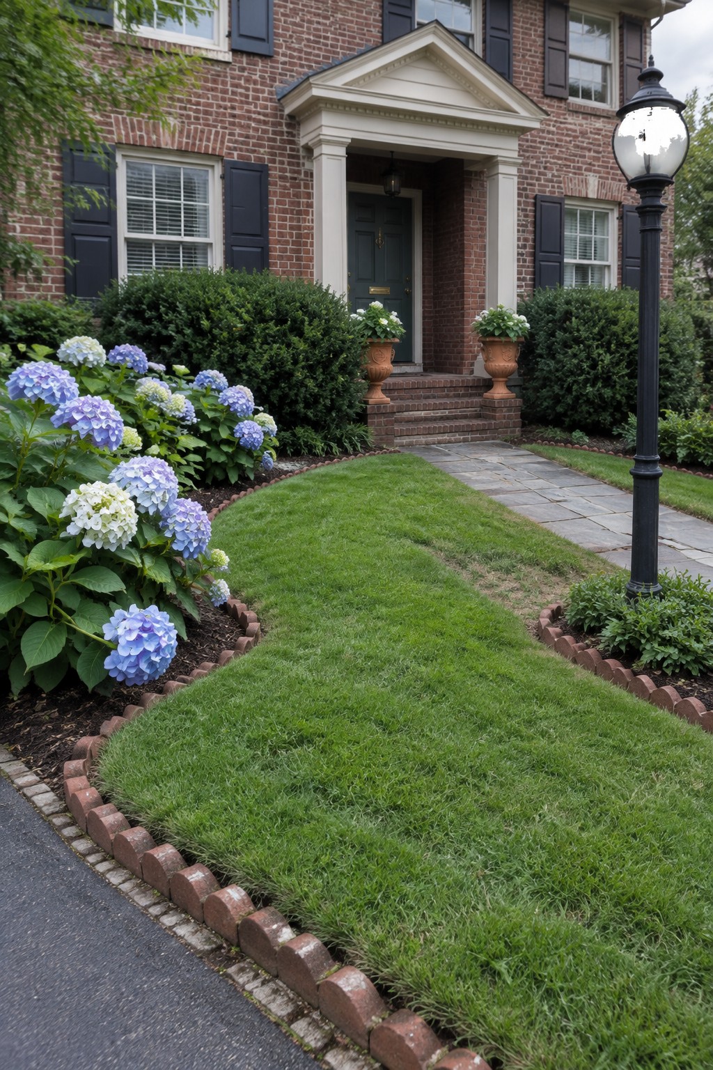 A front yard lawn bordered by curved red brick edging with blue and white hydrangea beds on the left and a stone path leading to a brick house entrance.