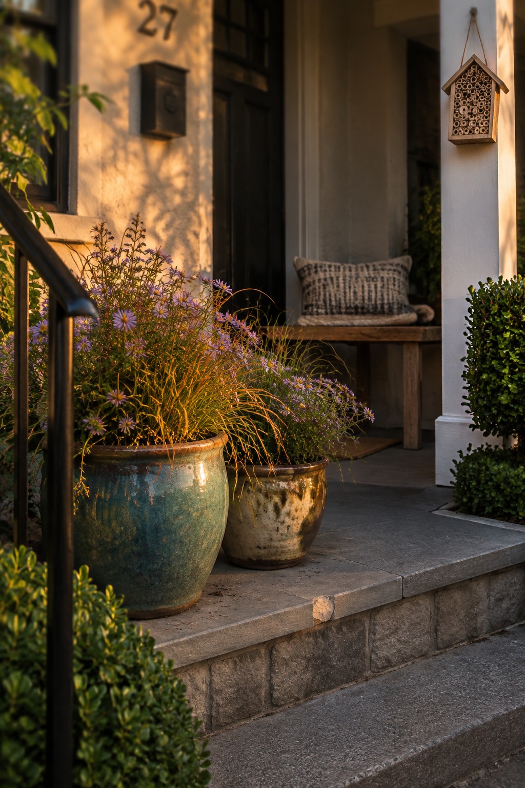 Two large ceramic pots with purple flowering plants sit on gray stone steps leading to a house entrance, with a wooden bench and insect hotel visible on the porch behind them.