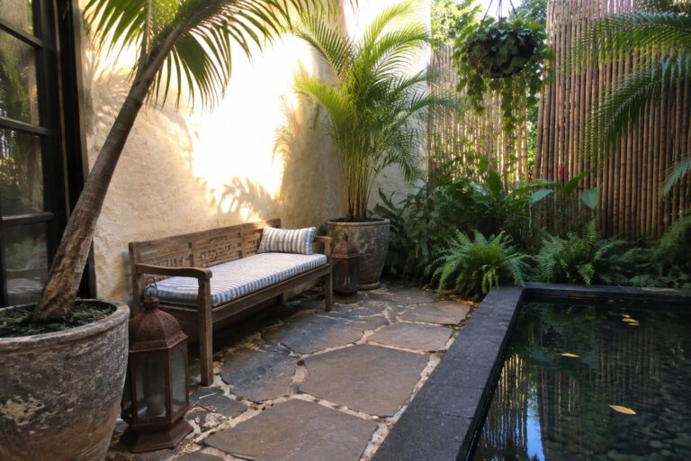 Lush tropical courtyard with stone path, wooden bench, potted palms, and reflecting pool