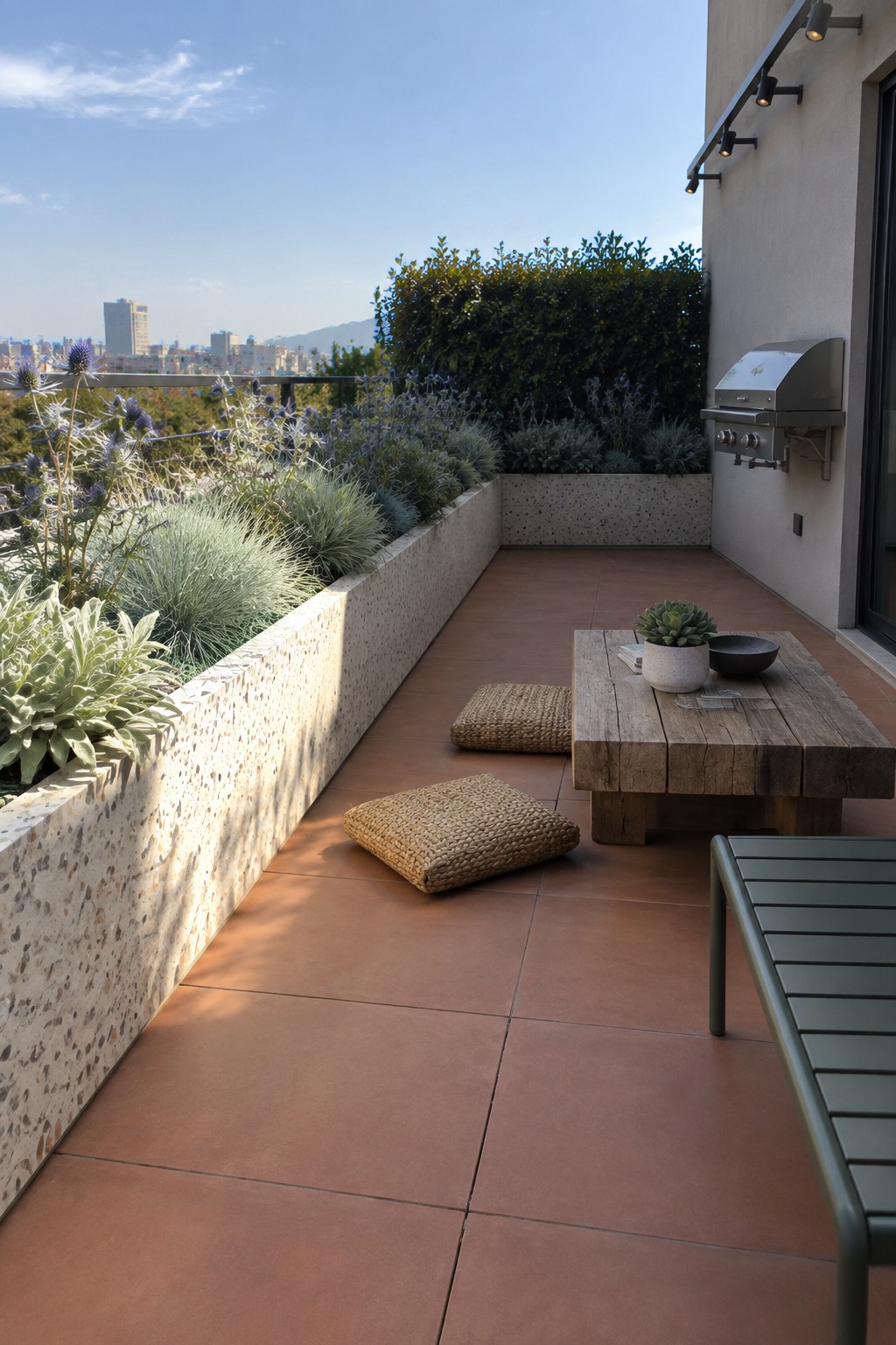 Rooftop terrace with long built-in concrete planters filled with grasses and flowering plants along one side, wooden table, woven floor cushions, and a wall-mounted grill.