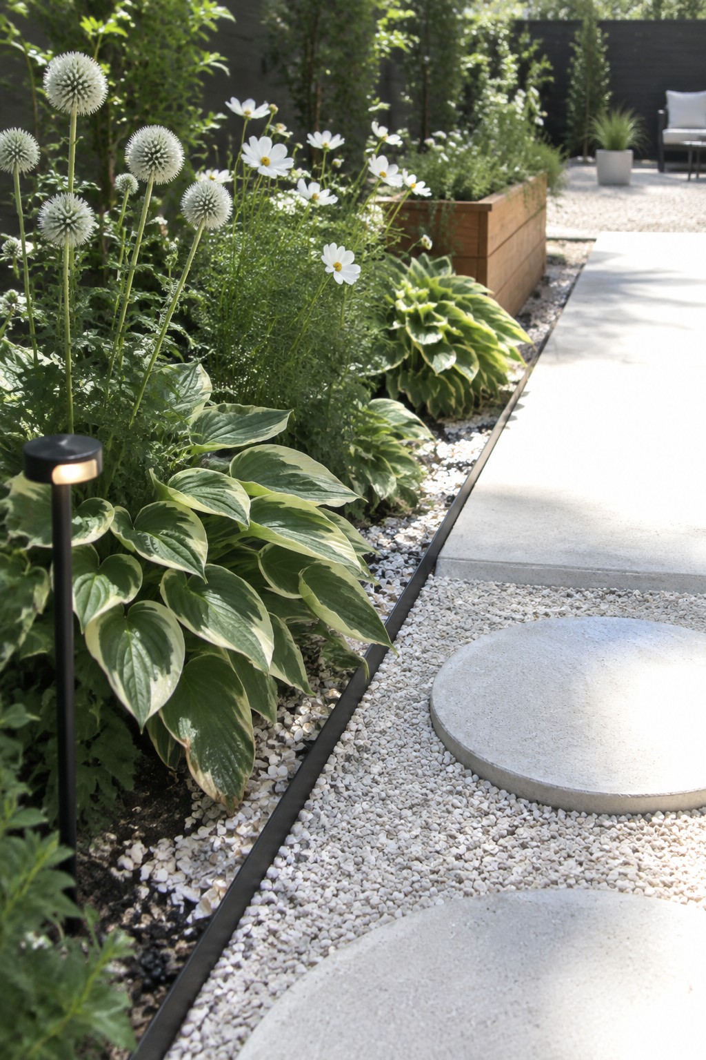 A garden bed with hostas, white flowers, and spherical blooms borders a gravel path with round concrete stepping stones.