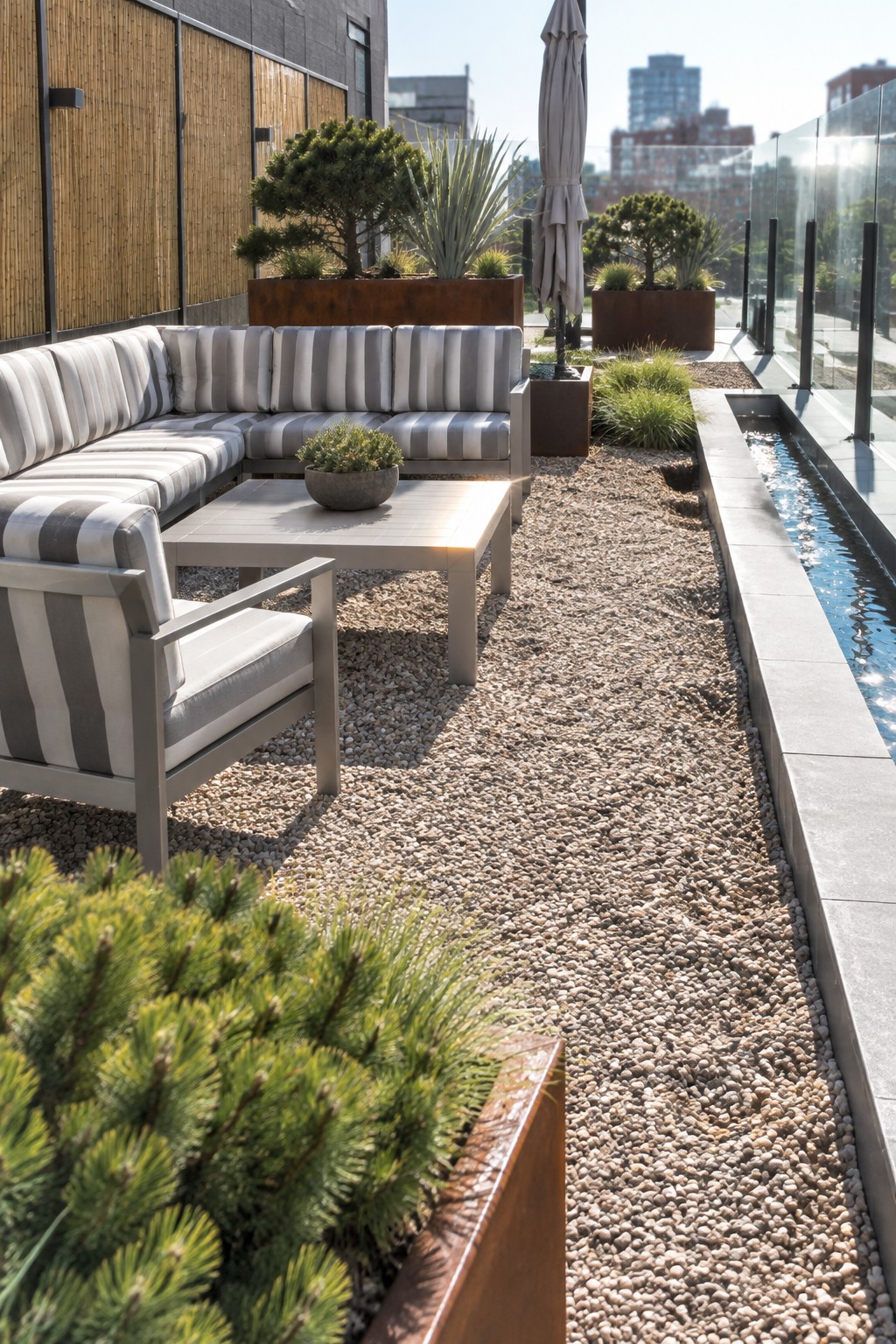 Modern rooftop garden with striped outdoor sectional sofa, gravel ground cover, water channel, and evergreen trees and shrubs planted in large rectangular metal containers.