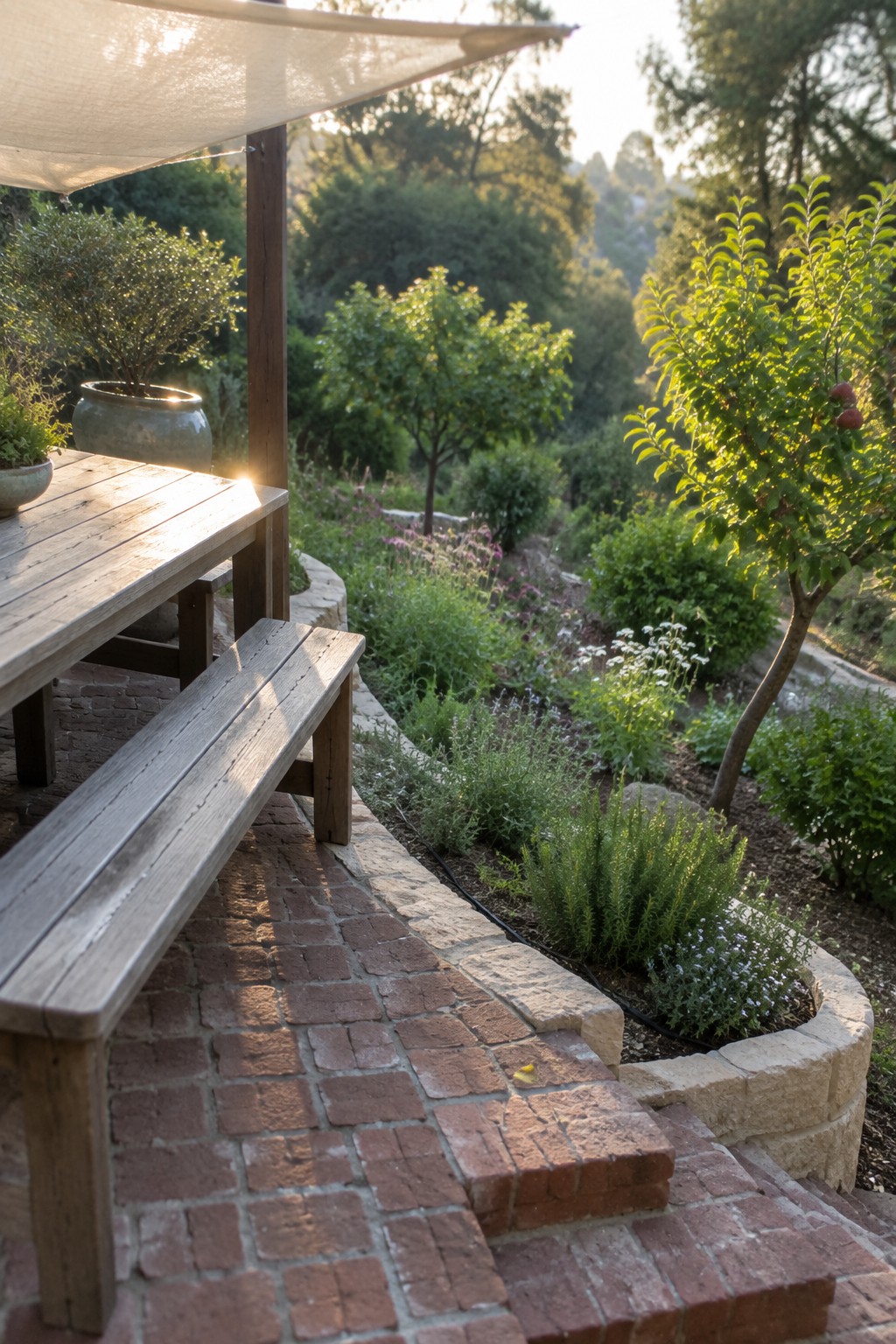 A wooden table and bench rest on a brick patio beside a curved stone-edged garden bed filled with herbs and shrubs, with trees and additional planting in the background.