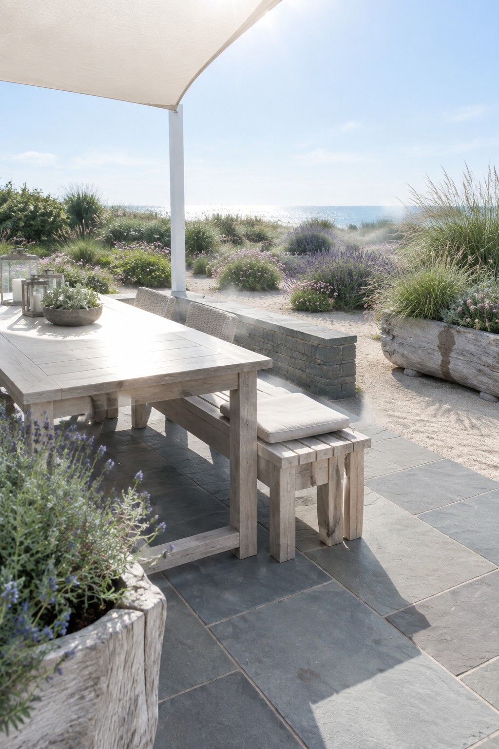 A light wood dining table and bench sit on a stone patio bordered by wooden planters filled with lavender and grasses, with a coastal garden and ocean beyond.