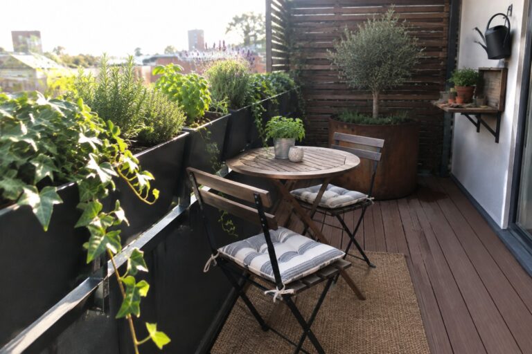 15 Smart Small Terrace Garden Ideas Using Railing Planters A narrow urban balcony with dark wood flooring, black railing-mounted planters filled with green plants and trailing ivy, a round wooden table with two folding chairs, and a large woven rug on the deck.