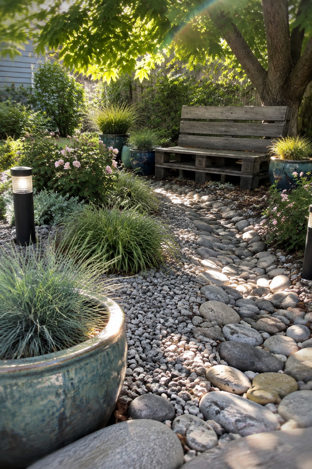 A garden path of small gray gravel mixed with larger smooth stones, bordered by ceramic pots holding grasses and flowers, with a wooden pallet bench in the background.