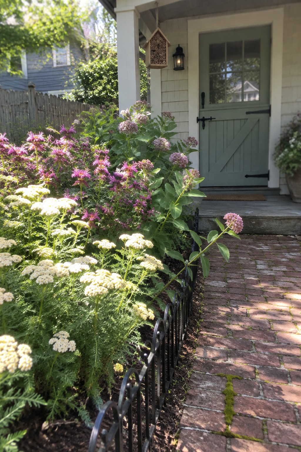 Flower bed with white yarrow and purple bee balm plants edged by a low black metal fence along a brick path beside a house with a green door.