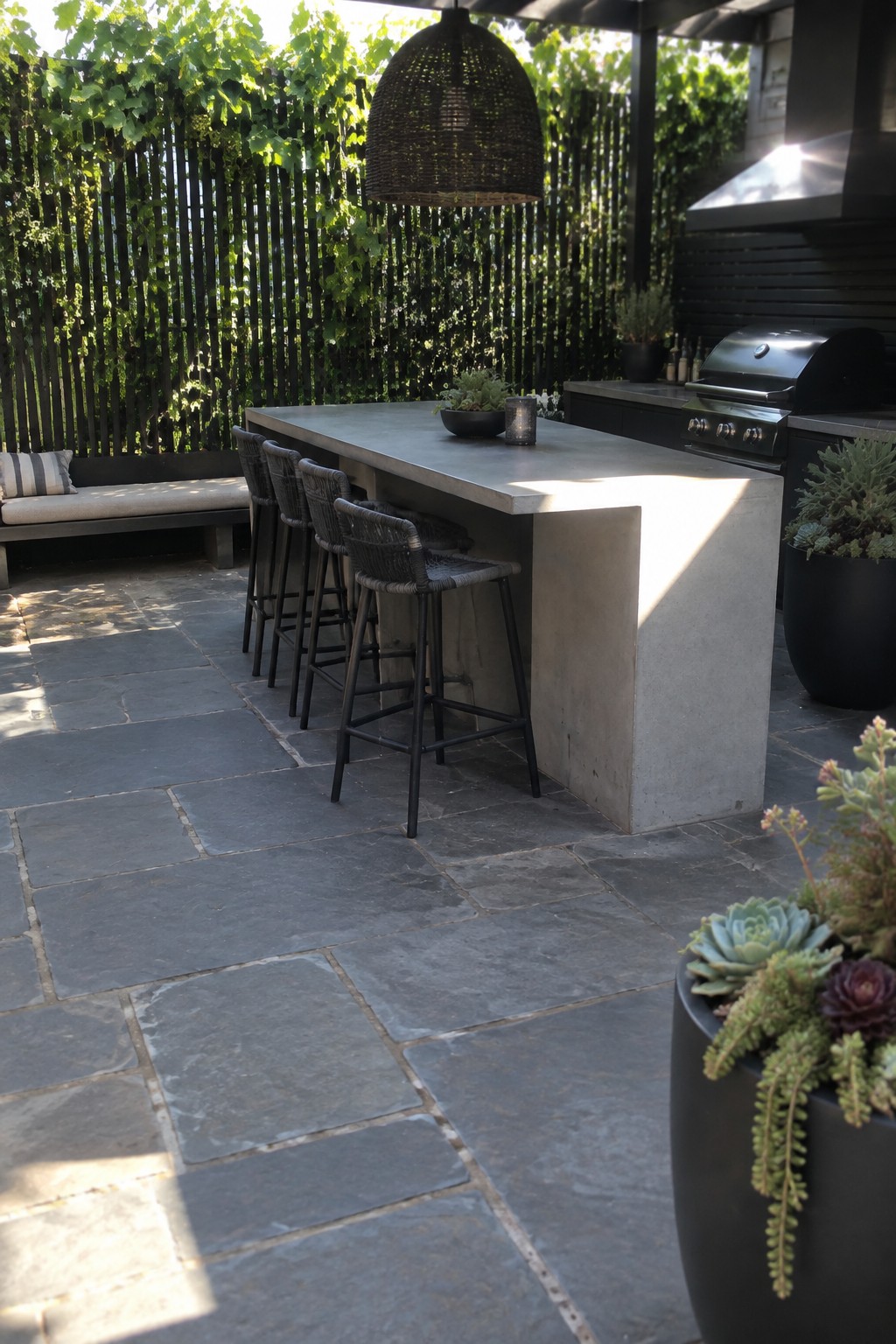 An outdoor concrete bar with woven barstools faces a dark wooden slatted fence covered in green climbing vines, with a grill and stone pavers visible nearby.