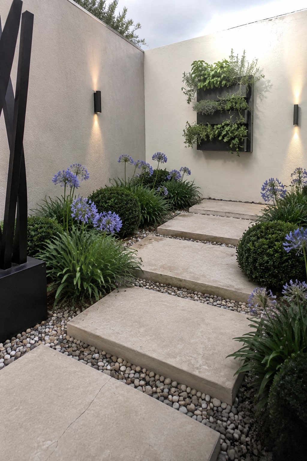 Large rectangular stone steps descend through a gravel path bordered by green shrubs and blue flowers, with a vertical planter on the wall.