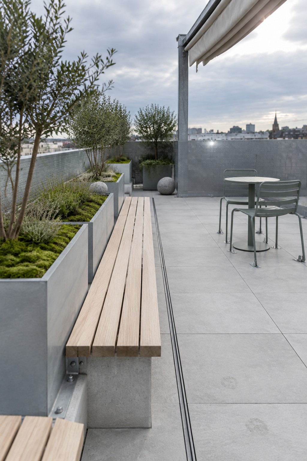 A rooftop terrace features large gray concrete planters with trees and green ground cover, a light wood bench attached to one planter, and a small table with chairs under an awning.
