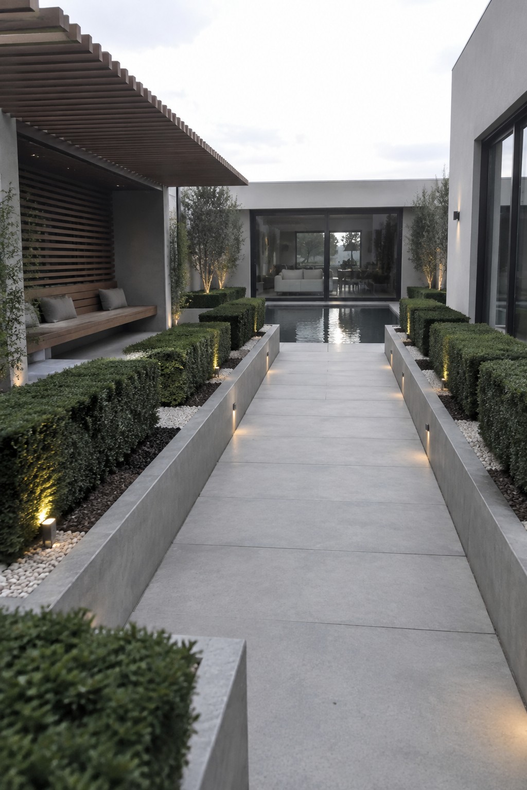 A straight concrete walkway bordered by long rectangular planters filled with uniform green hedges and small ground lights.
