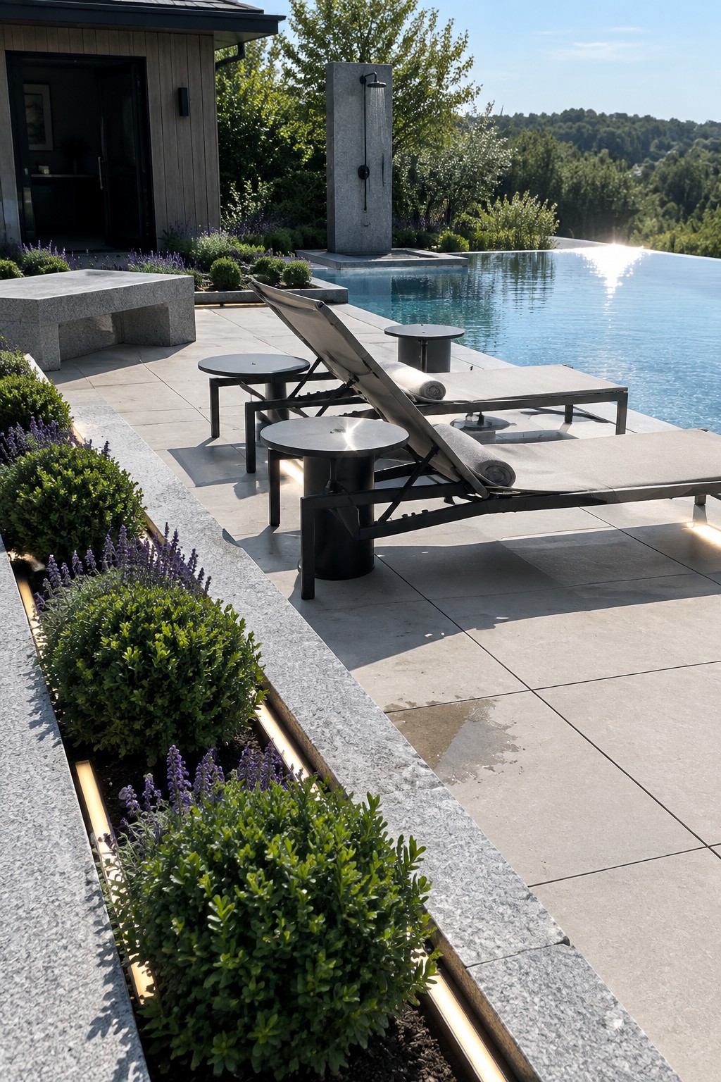 A modern stone patio with lounge chairs next to an infinity pool, bordered by a low natural stone edge and planted beds containing green shrubs and purple flowers.
