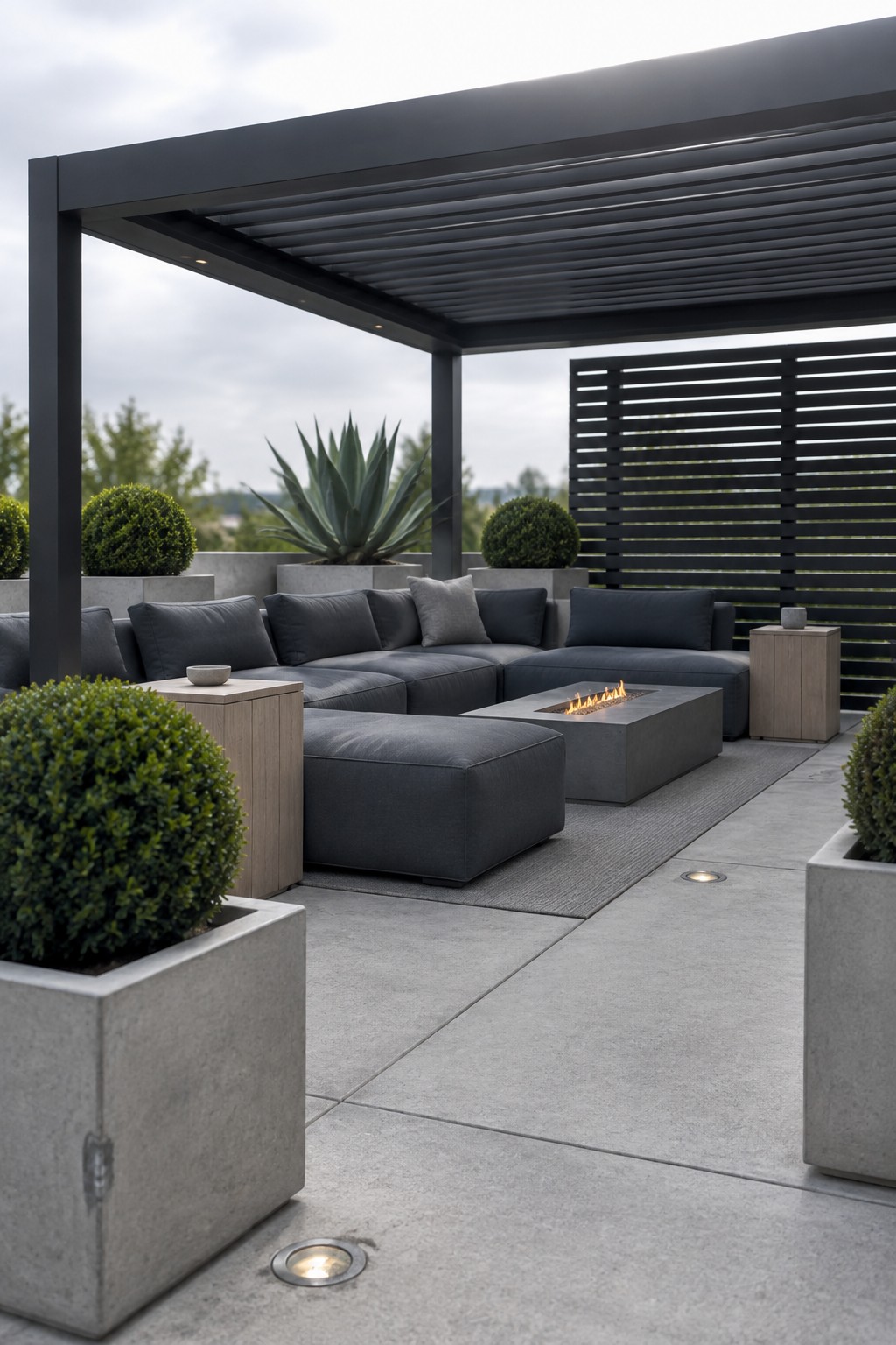 A modern outdoor patio under a dark metal pergola with horizontal slats, featuring a gray sectional sofa arranged around a rectangular concrete fire pit on a paved surface with potted shrubs.