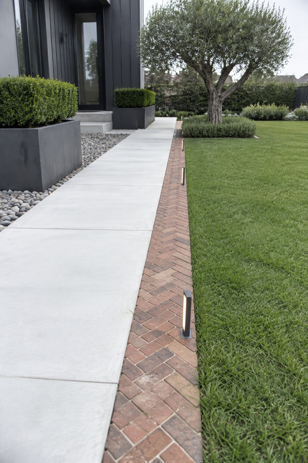 A straight concrete walkway bordered by herringbone brick edging with small integrated lights, running beside a green lawn in front of a modern house.