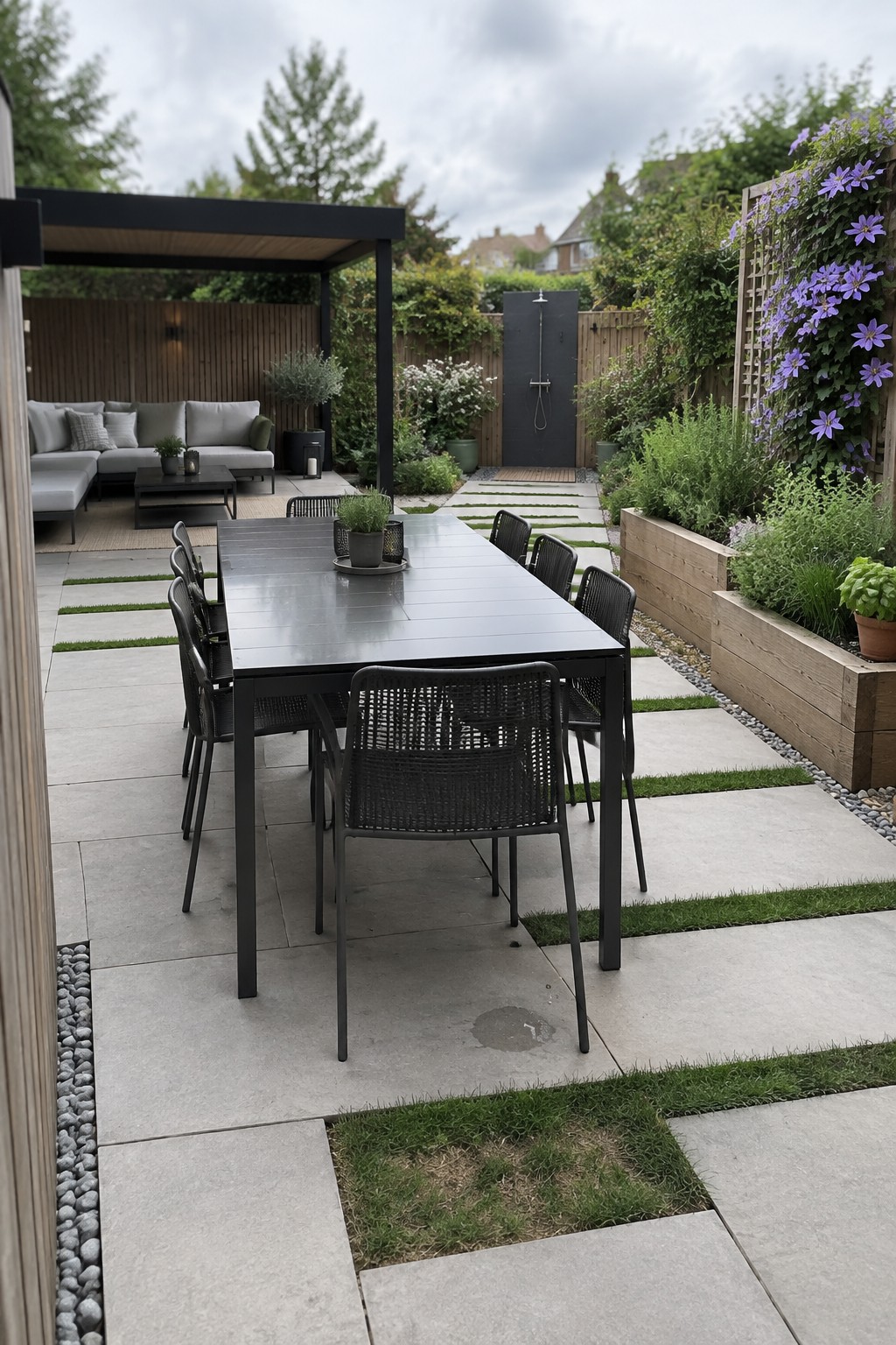 A backyard patio featuring a large outdoor dining table with black chairs, a covered lounge area under a pergola, an outdoor shower, and raised garden beds along the sides.