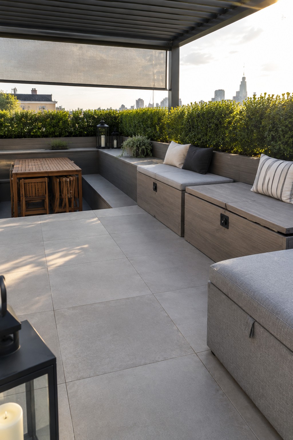 Rooftop terrace with built-in wooden storage benches topped with gray cushions, a wooden table with folded chairs, and greenery along the perimeter.
