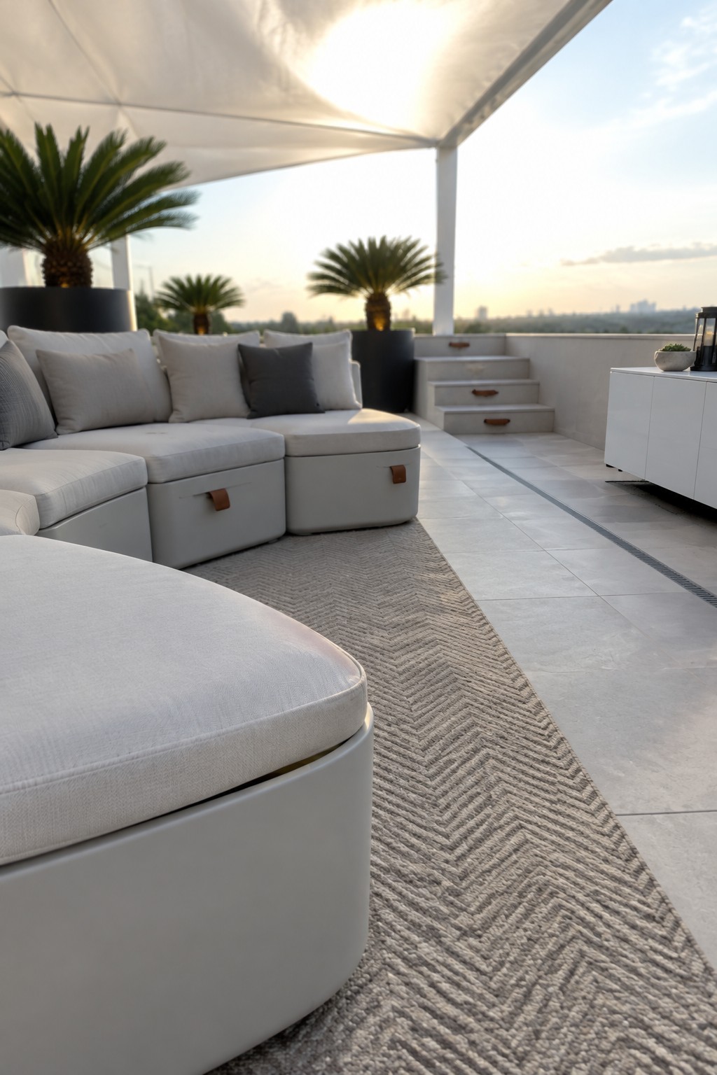 A rooftop terrace shows a large curved sectional sofa in light gray fabric with integrated storage compartments under the seats, placed on a patterned rug beneath a white shade canopy.