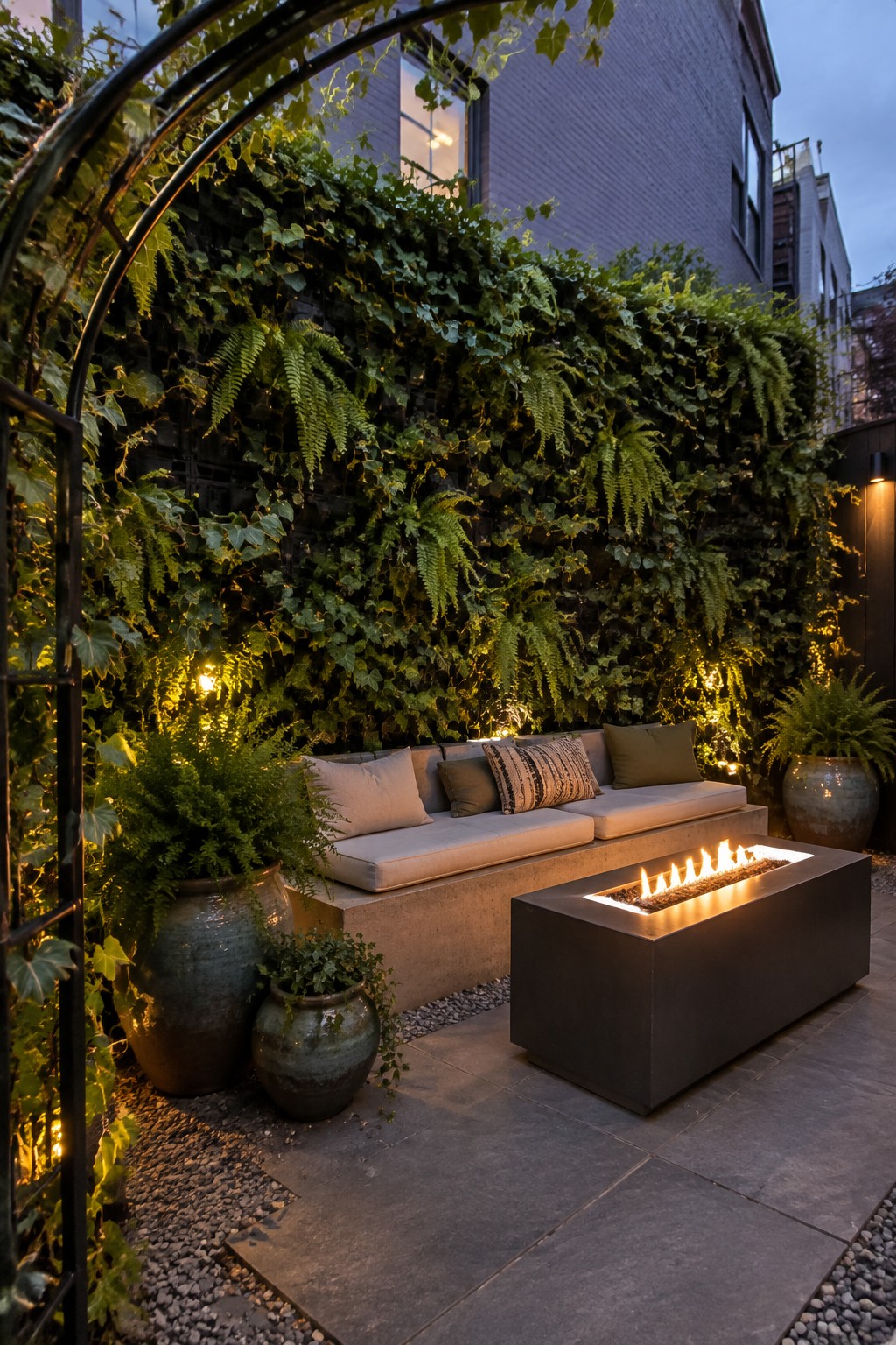 A small outdoor patio with a dense living wall of ferns and ivy, a built-in bench with cushions, and a rectangular fire pit on stone paving.