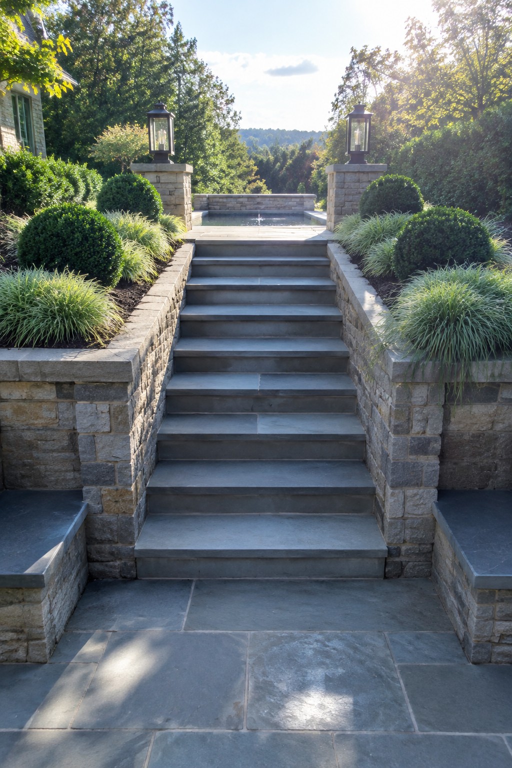 A wide stone staircase rises between matching stone walls topped with rounded shrubs, ornamental grasses, and lantern-topped pillars, with a small fountain visible at the top.