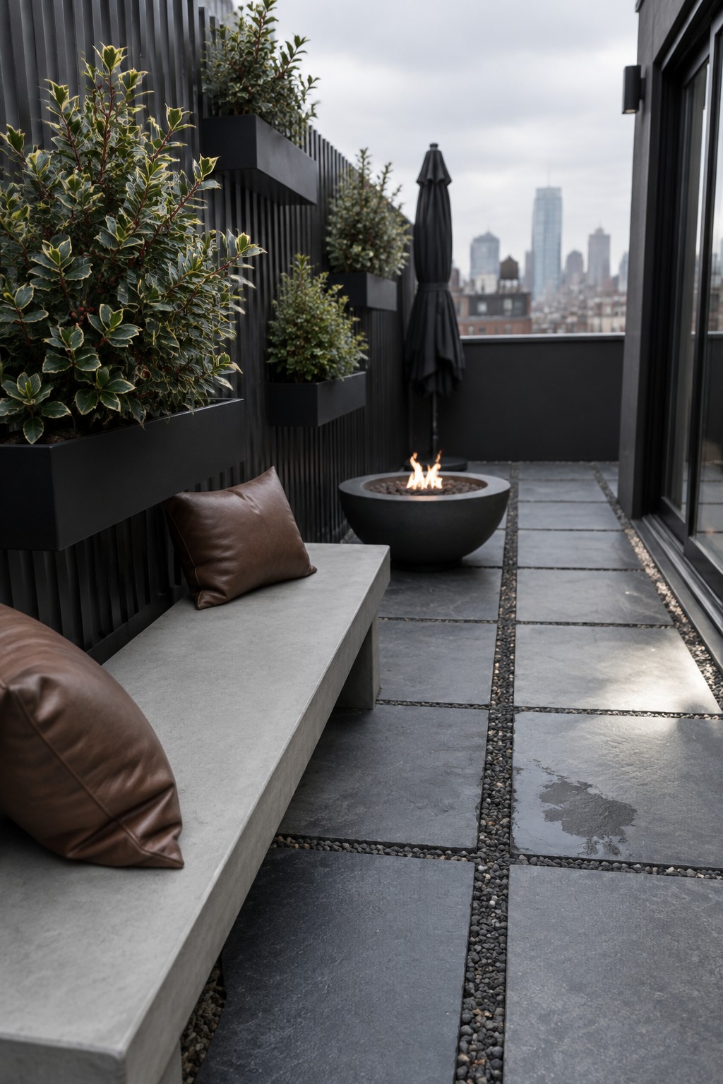 Rooftop terrace with concrete bench, brown cushions, black slatted wall with mounted evergreen planters, round fire pit on dark pavers, and city skyline beyond.