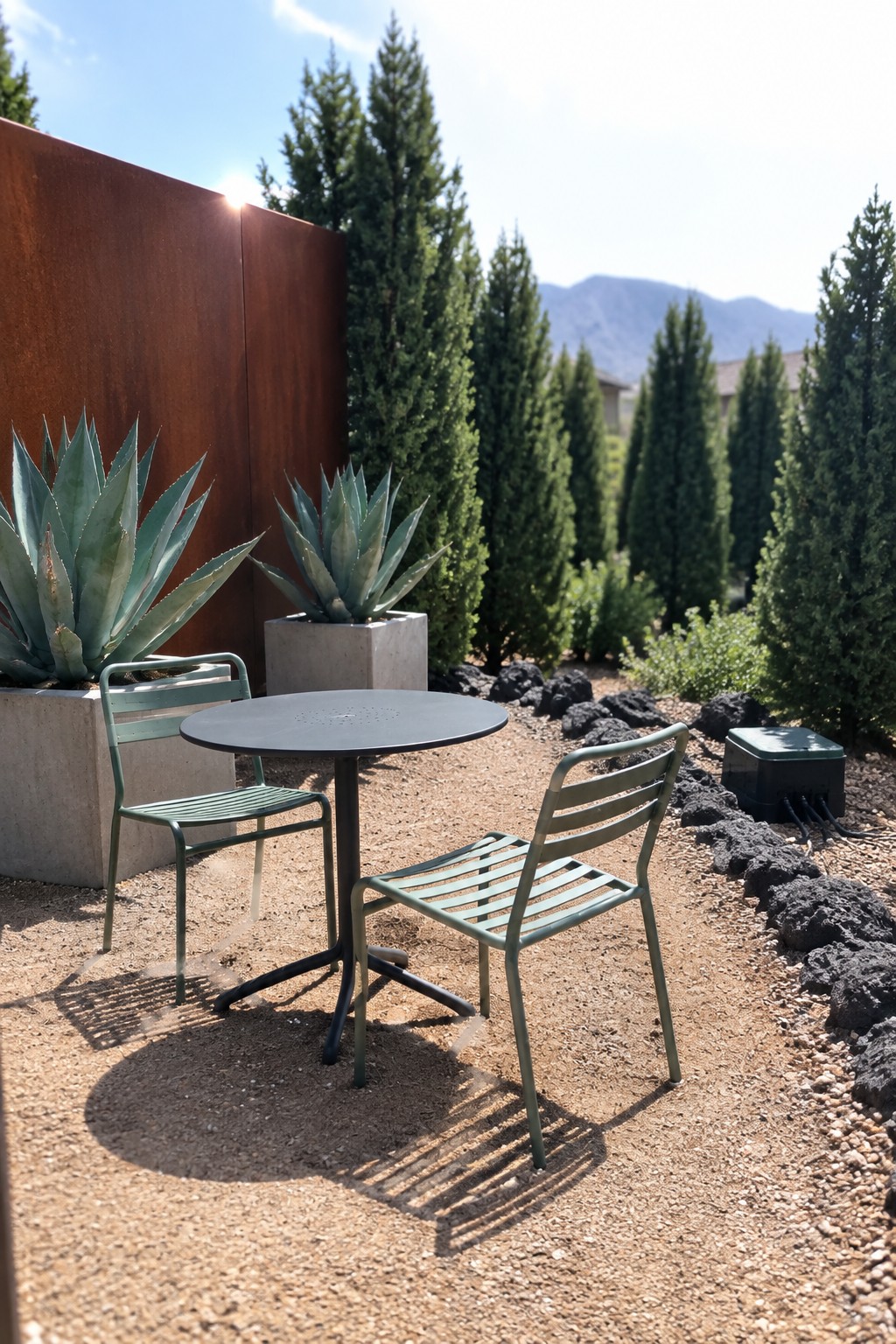 A round black table with two green metal chairs sits on gravel next to a rusted metal wall, large potted agave plants, and tall evergreen trees.