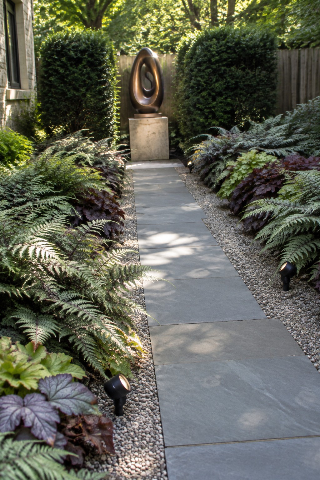 A narrow stone path runs between beds of ferns and other shade plants, with small lights along the gravel edges and a bronze sculpture visible at the end.