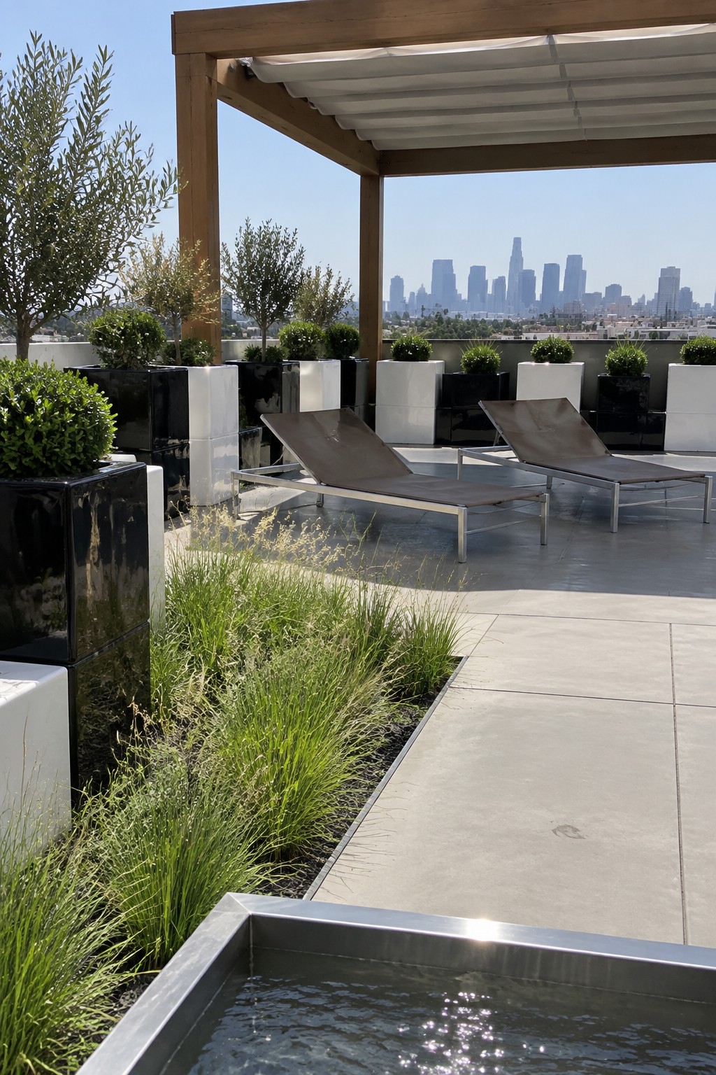 A rooftop terrace with two brown lounge chairs, black and white geometric planters, a metal water feature, and a distant city skyline.