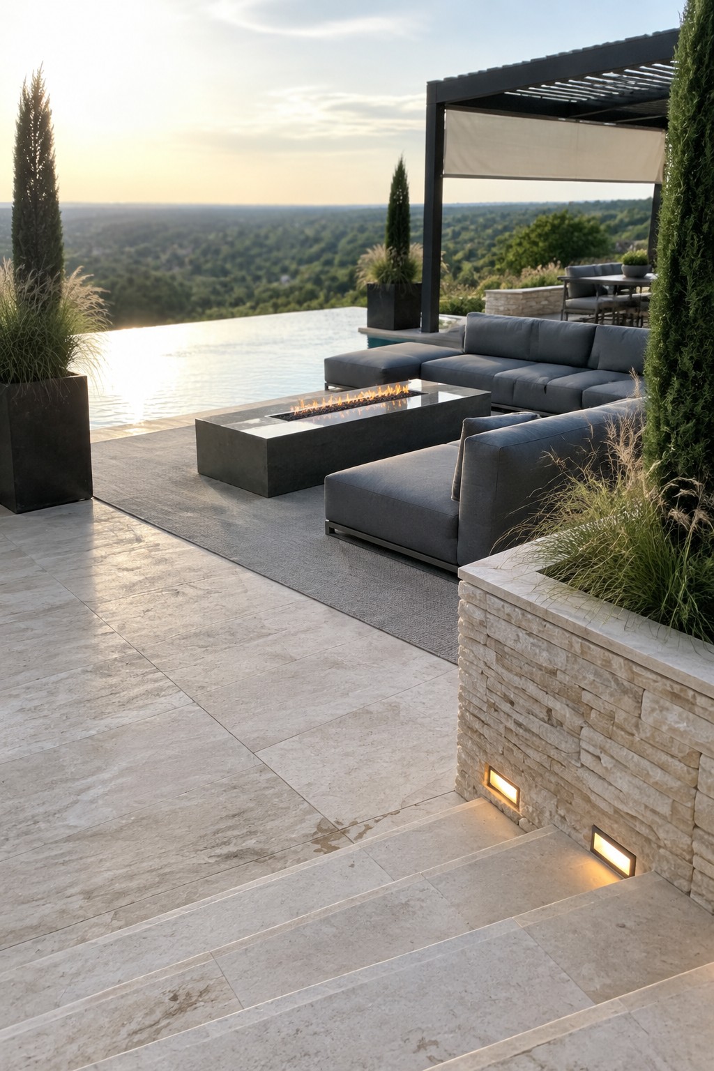 A modern outdoor patio features a gray sectional sofa arranged around a rectangular fire pit beside an infinity pool, with stone steps and planters in the foreground.