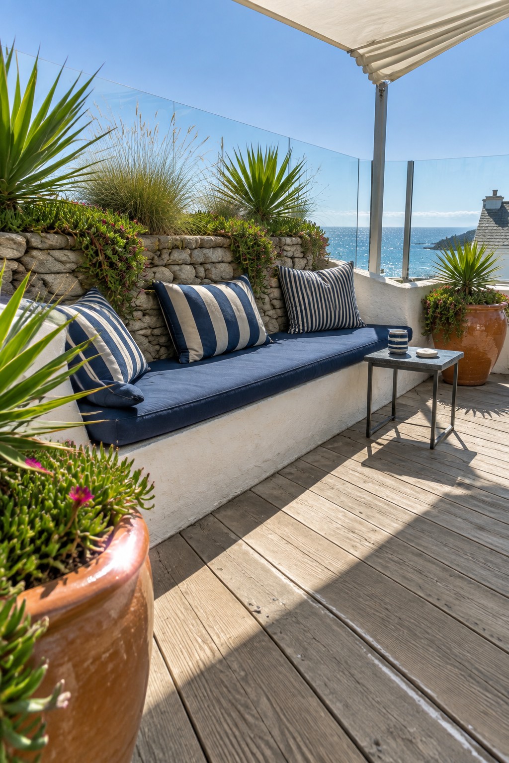 A roof terrace featuring a built-in bench with blue cushions along a stone wall topped with plants, a small side table, wooden decking, and an ocean view.