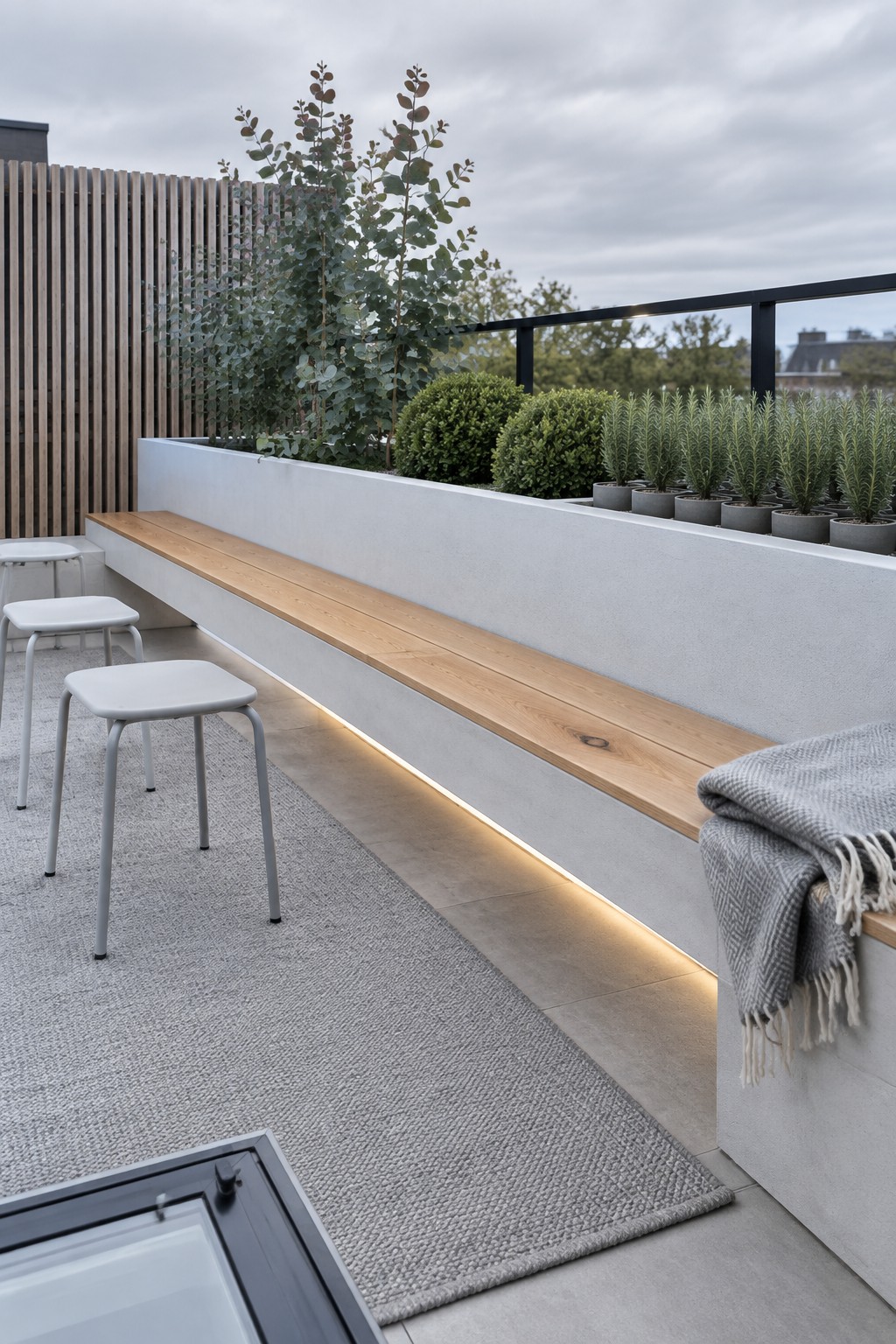A modern roof terrace shows a long wooden bench built into a white concrete planter wall filled with green plants and small trees, with three gray stools on a woven rug nearby.